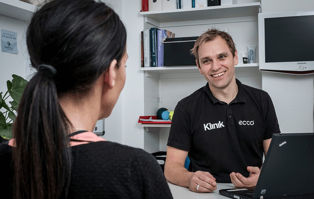 Smilende behandler i sort polo taler med en kvindelig patient ved skrivebordet
