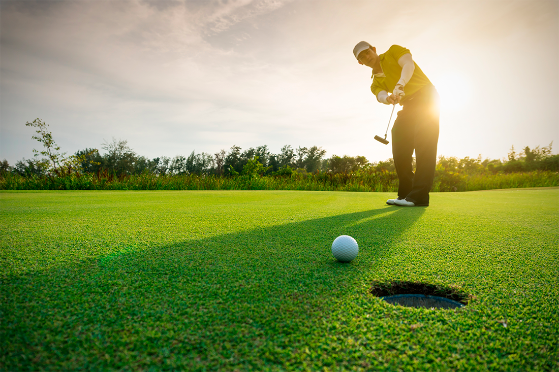 Golfspiller med hvid kasket i putterpositur på green i solnedgang