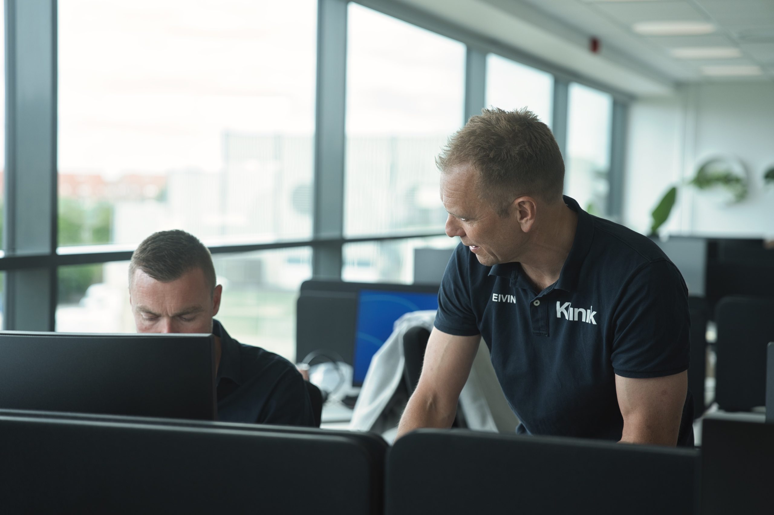 Mand i Klinik-polo læner sig ind over kollegaens laptop ved skrivebordet